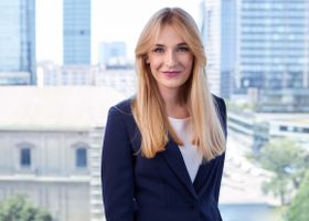 Smiling blonde businesswoman in elegant attire standing against a Warsaw urban backdrop.